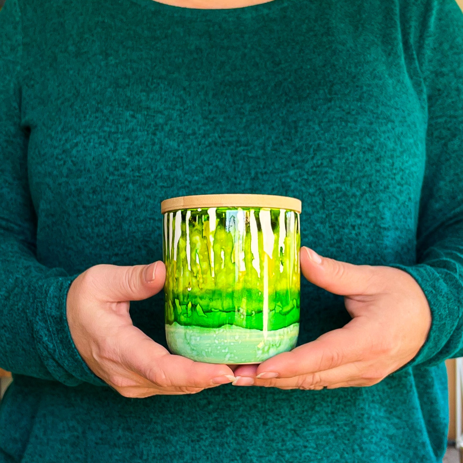 Holding a tall, two wick ceramic soy candle handpainted in layers of mottled greens