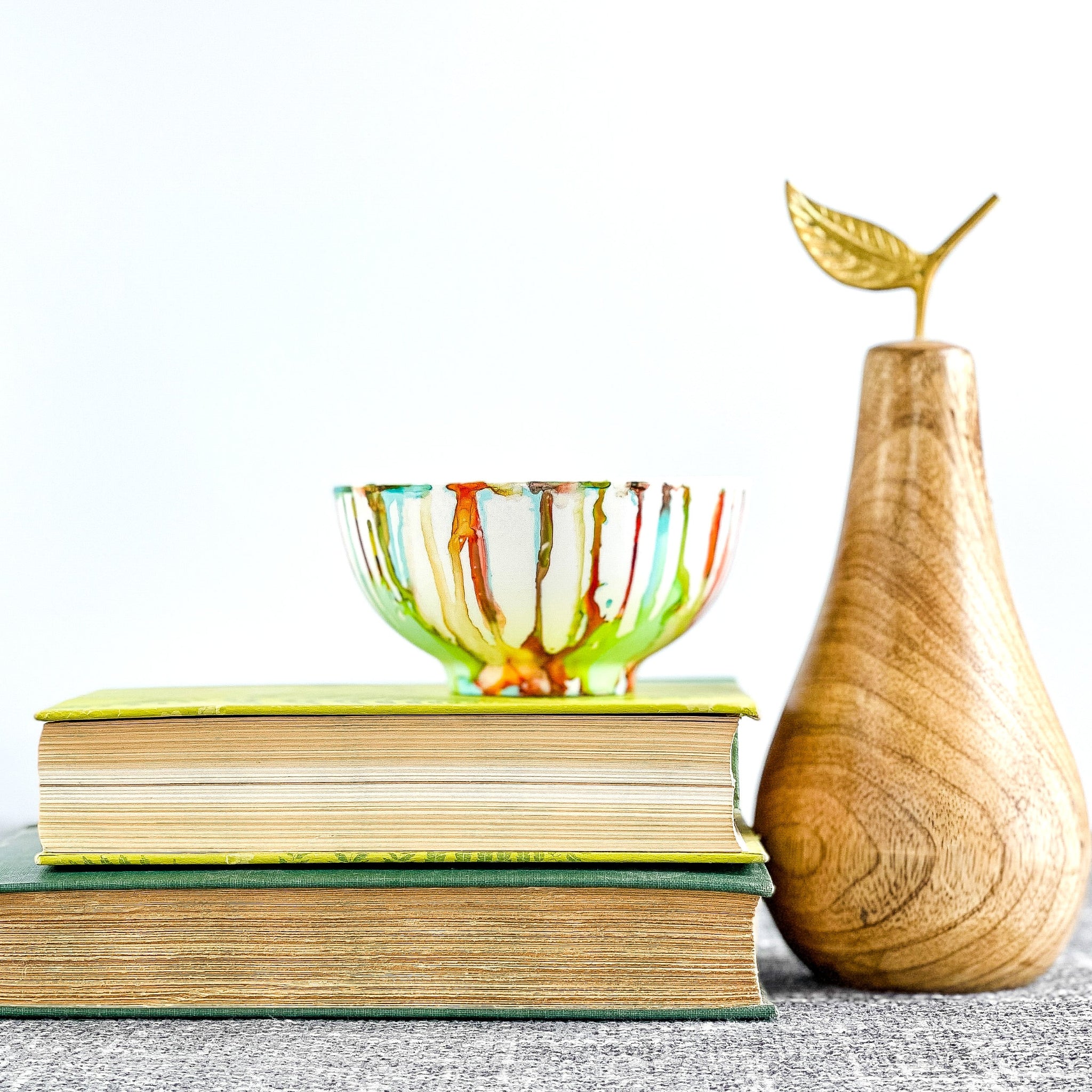 A hand-painted multicolored candle vessel with vibrant shades of green, orange, red, and teal, displayed on a stack of books next to a wooden pear.