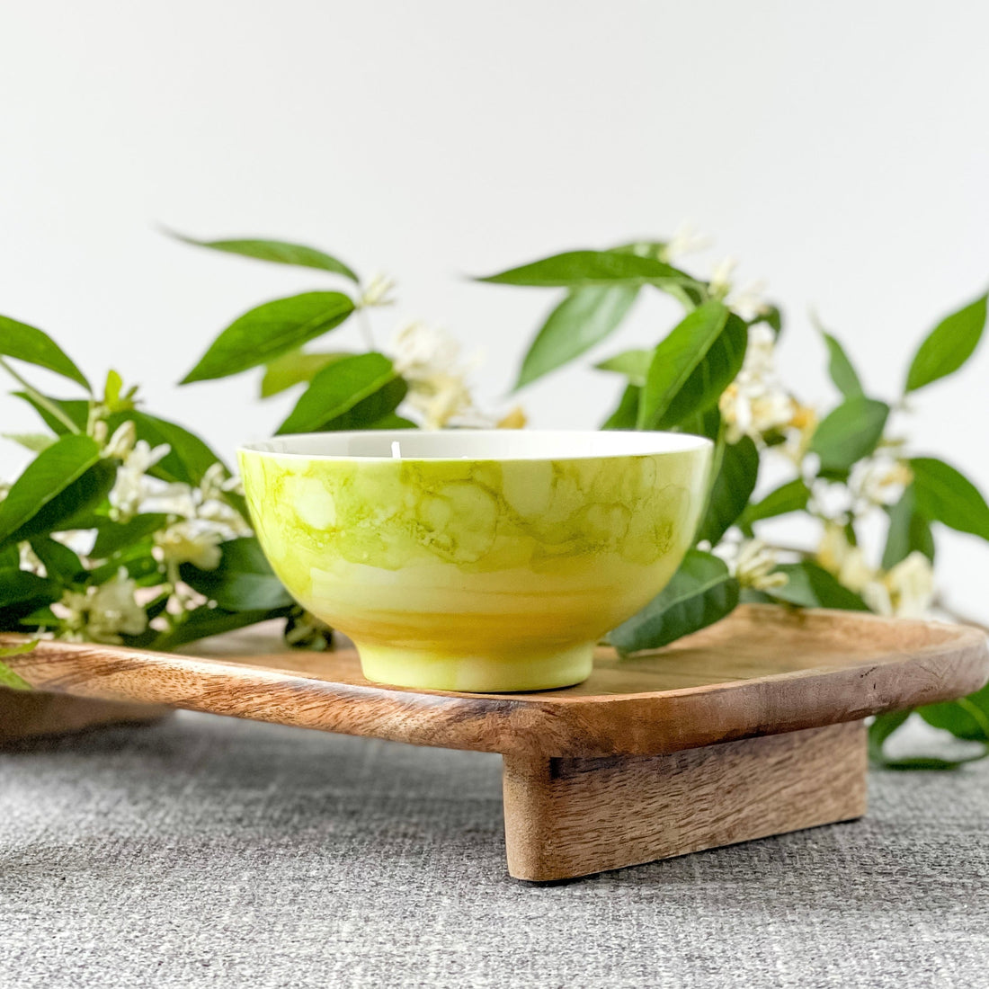 A ceramic candle with a triple wicked design, hand painted in mottled mossy greens, displayed on a wooden tray with green foliage in the background.