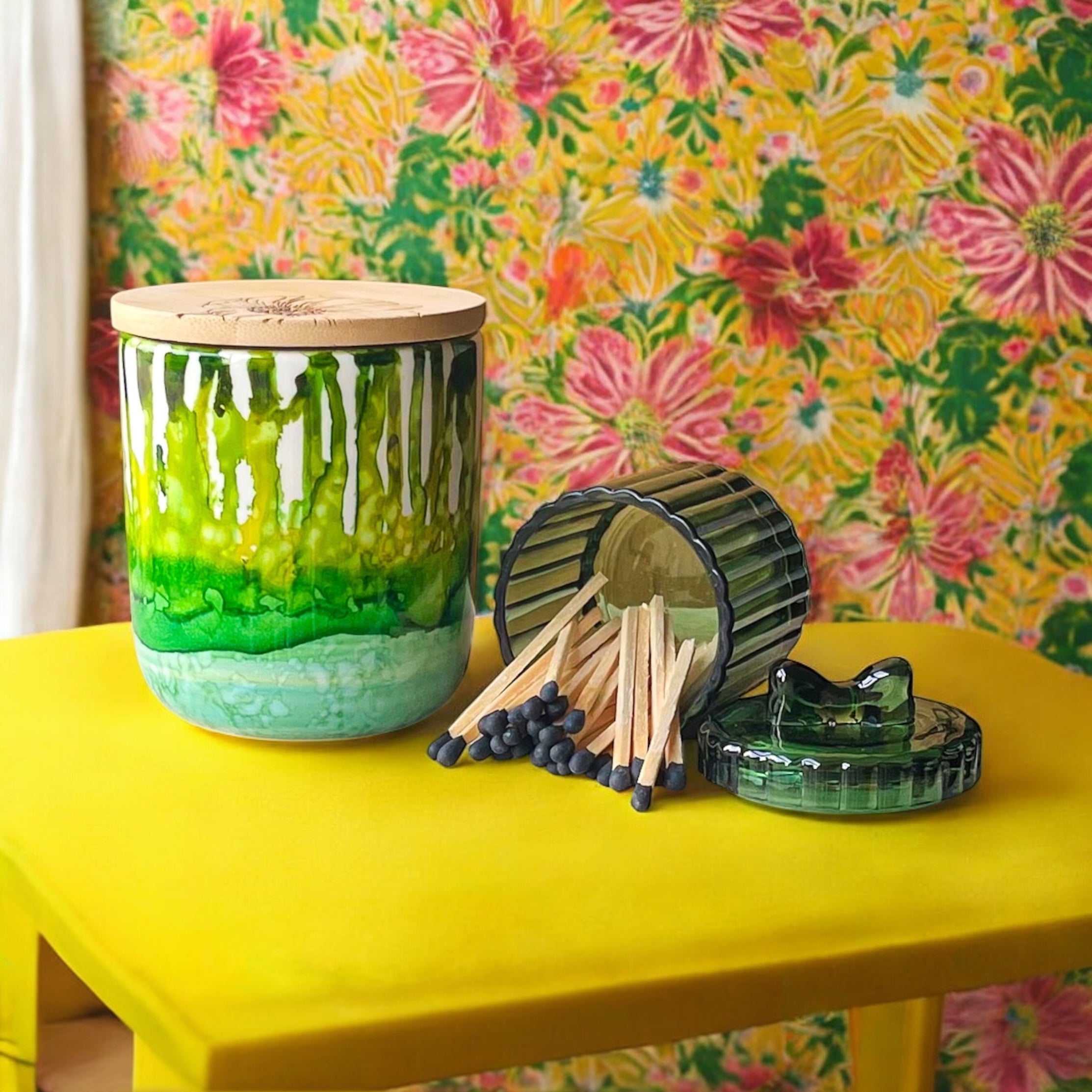 Tall, two wick ceramic soy candle handpainted in layers of mottled greens on a yellow table with matches