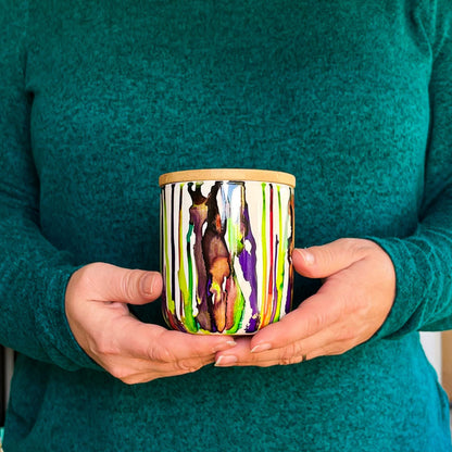Hands holding a tall, two wick ceramic soy candle handpainted in bright greens and deep purples