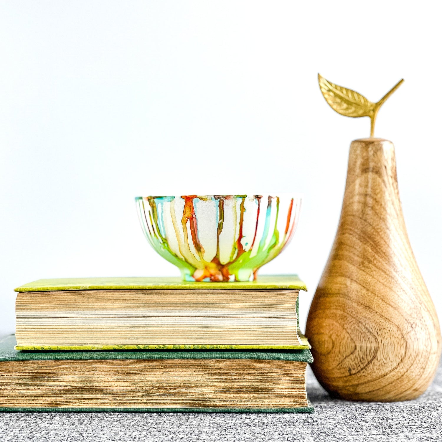 A hand-painted multicolored candle vessel with vibrant shades of green, orange, red, and teal, displayed on a stack of books next to a wooden pear.