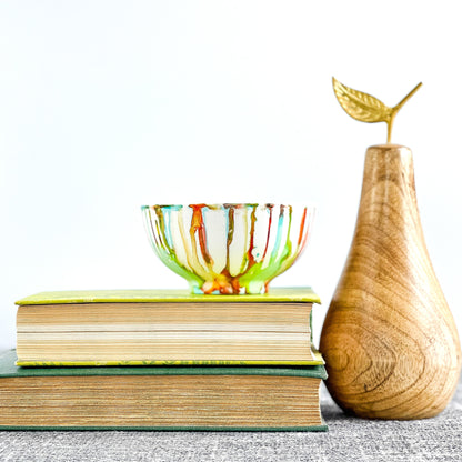 A hand-painted multicolored candle vessel with vibrant shades of green, orange, red, and teal, displayed on a stack of books next to a wooden pear.