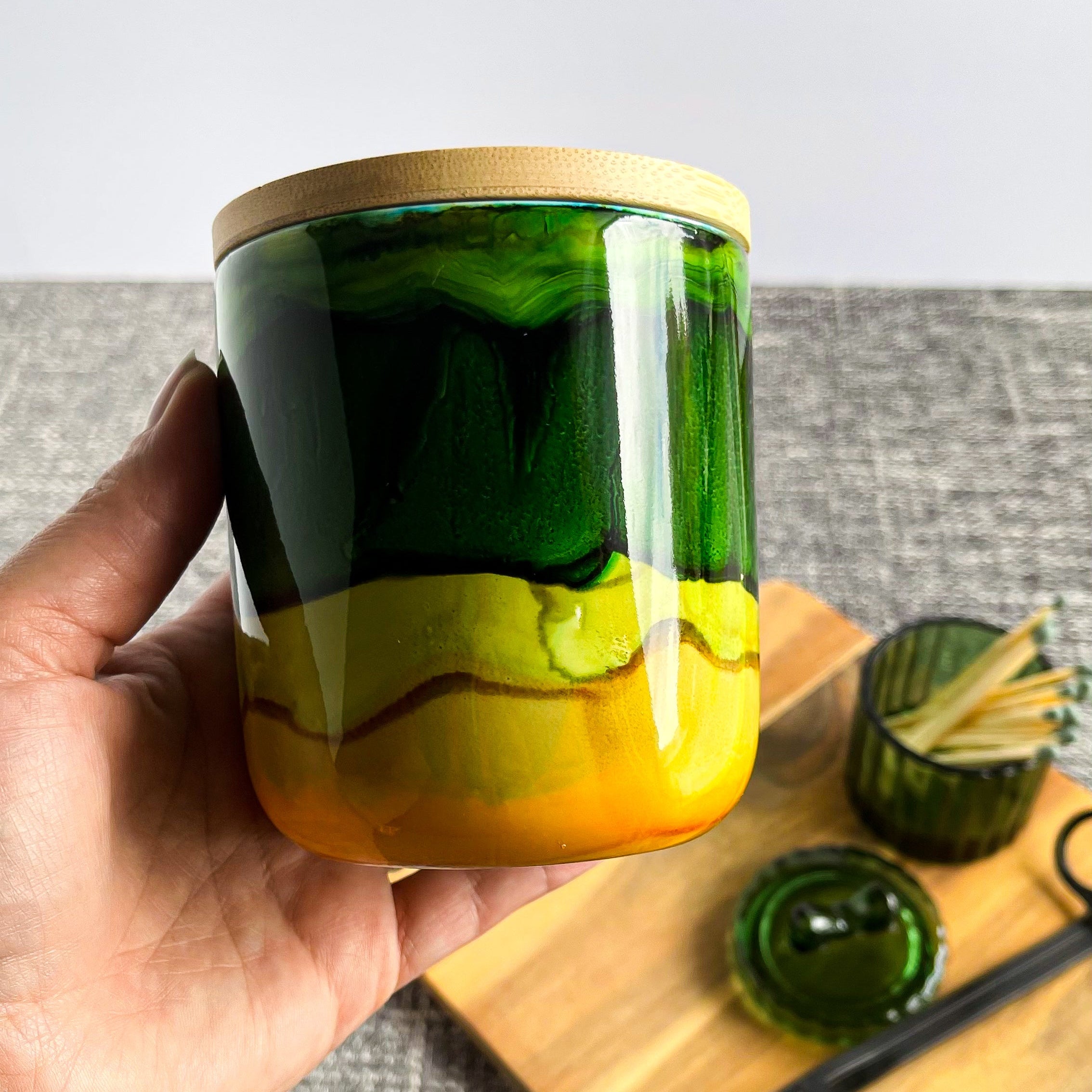 Hand holding a tall, two wick ceramic candle handpainted in bands of mossy and forest greens with a base of warm yellow